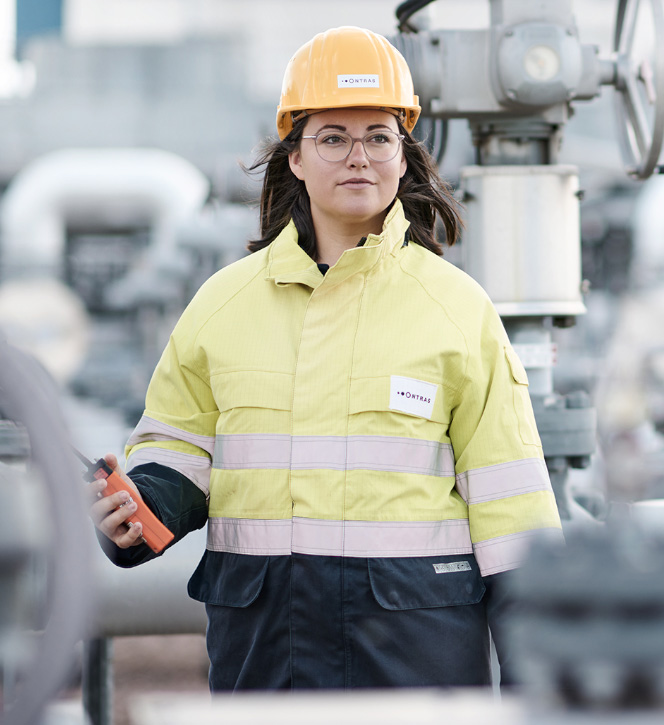 Eine junge ONTRAS-Mitarbeiterin steht auf einer Betriebsanlage. Sie trägt gelbe Arbeitsschutzkleidung und einen gelben Helm. Sie blickt in die Ferne. In der Hand hält sie ein Gasspür-Gerät.