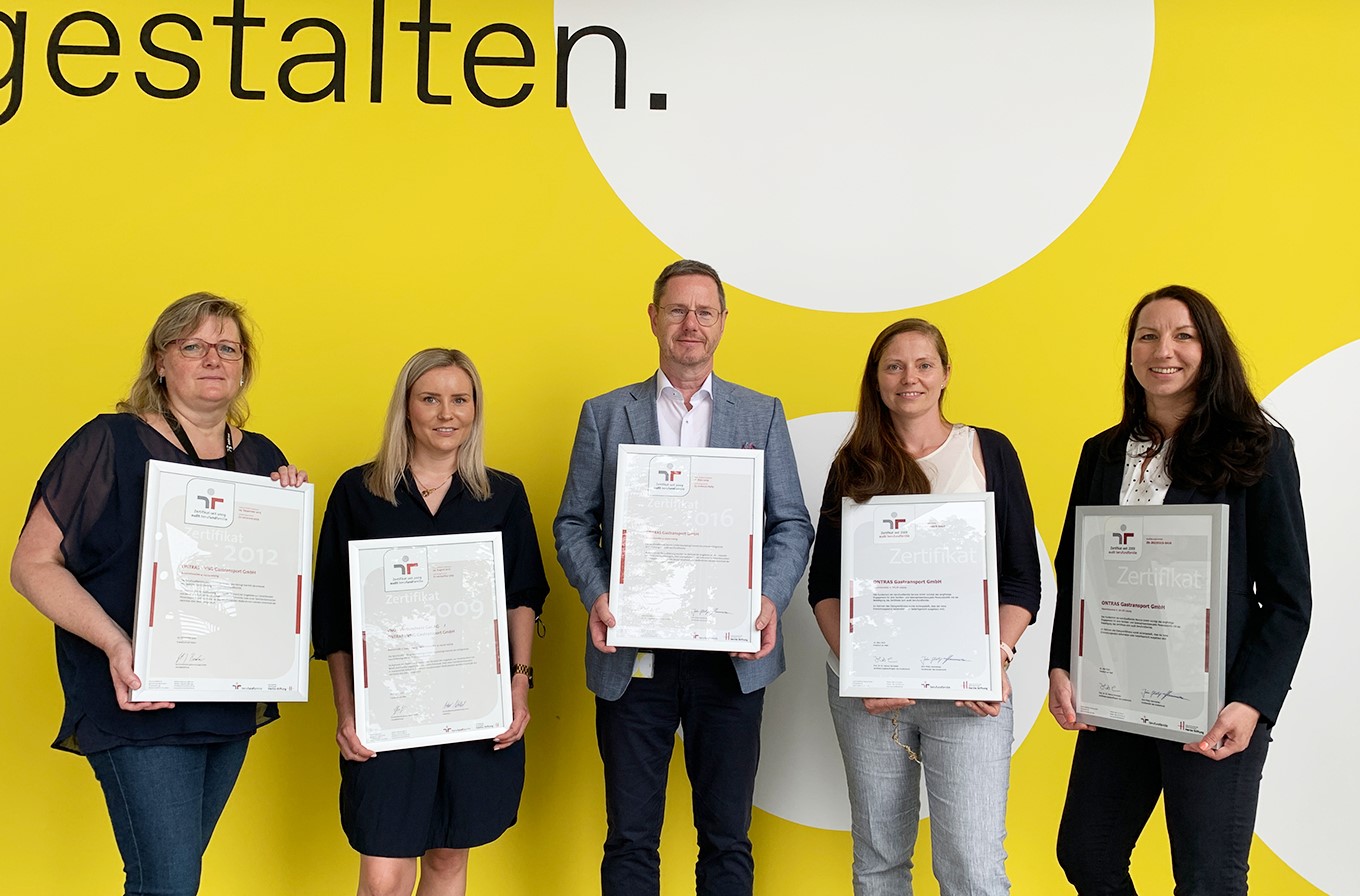 Lächelnde Mitarbeitende von ONTRAS zeigen die fünf Zertifikate des erfolgreichen Audits berufundfamilie vor einer gelben Wand im ONTRAS-Design
