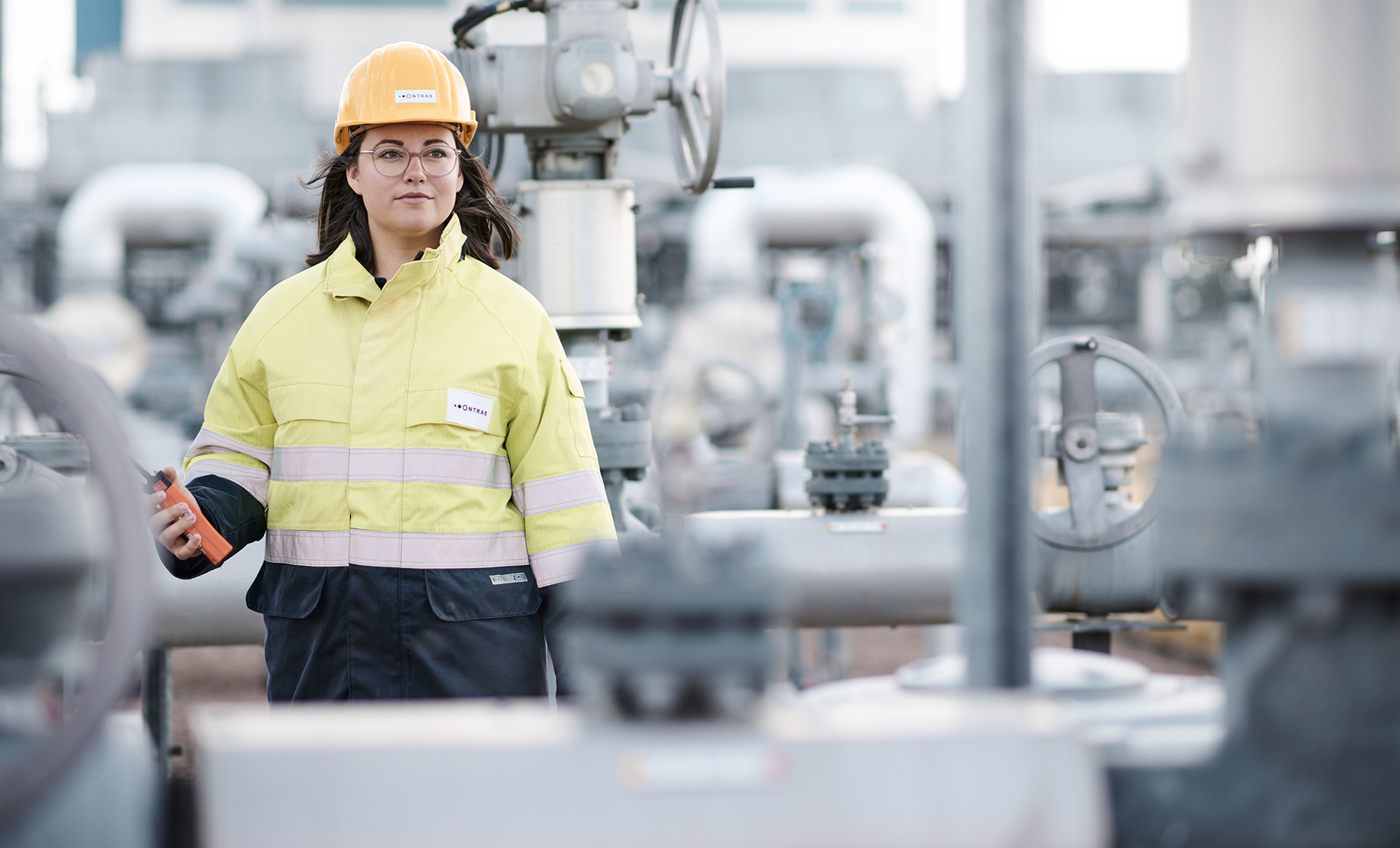 Eine junge ONTRAS-Mitarbeiterin steht auf einer Betriebsanlage. Sie trägt gelbe Arbeitsschutzkleidung und einen gelben Helm. Sie blickt in die Ferne. In der Hand hält sie ein Gasspür-Gerät.